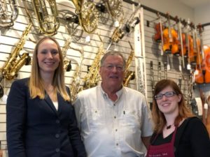 A tall young blonde white woman, an average height middle aged white man with glasses, and a short white red-headed woman with glasses stand in front of a wall of instruments.