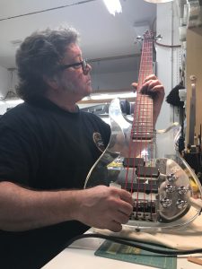 A white man stands at a shop bench and fingers a clear plexiglass bass guitar while checking string tension. The bass has red strings, skull knobs with red crystal eyes, and custom pickups.