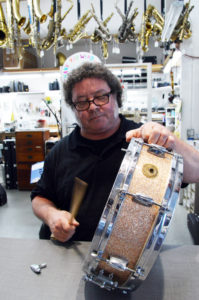 A white man of middle age stands at a shop counter and taps a drum stick on the top head of a champagne colored snare drum that he holds on its side.