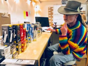 A white man with dreadlocks, a fedora, and a rainbow sweater sits at a bench, contemplating several boxes of Vandoren reeds.