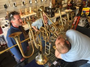 Three people, two men and one woman, are gathered around a complicated looking brass sculpture with seven bells. The taller of the two men is attempting to play one of the seven mouthpieces that emerge from it.