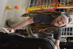 A man in a warehouse with long hair kneels behind a tuba case while wiping it down with a small cloth.